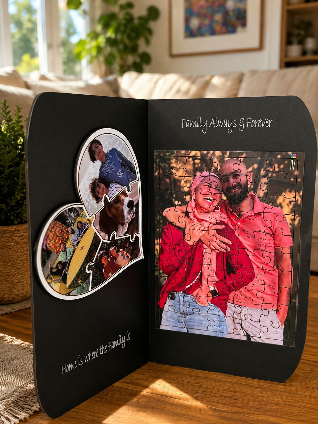 A black personalized jigsaw puzzle display stand shown open on a wooden table in a living room. The left panel has a heart-shaped collage of family photos with the text 'Home is where the family is,' and the right panel shows a completed puzzle insert with the text 'Family Always & Forever.